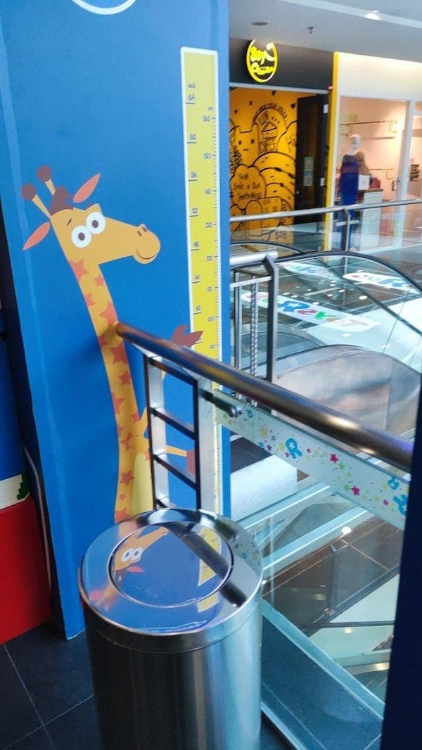 Giraffe holding a ruler to measure kids was installed on the wrong wall at the mall, so the ruler is on the wrong side of a barrier, where there is a drop to the floor below.